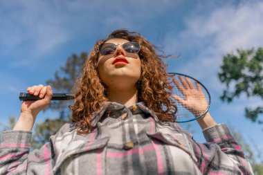 Ağaçlarla kaplı bir parkta genç, kıvırcık esmer bir kız güneşli bir günde elinde bambinton bir raketle kameraya poz veriyor.