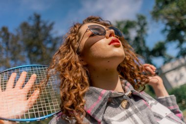 Ağaçlarla kaplı bir parkta genç, kıvırcık esmer bir kız güneşli bir günde elinde bambinton bir raketle kameraya poz veriyor.