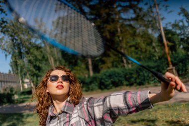 Ağaçlarla kaplı bir parkta genç, kıvırcık esmer bir kız güneşli bir günde elinde bambinton bir raketle kameraya poz veriyor.