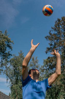 Ağaçlarla çevrili bir parkta, mavi tişörtlü, güneş gözlüklü esmer bir adam oyun başlamadan önce voleybol topunu ısıtıyor ve sokakta topla poz veriyor.