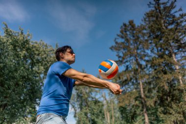 Dışarıda ağaçlarla dolu bir parkta mavi tişörtlü ve güneş gözlüklü esmer bir adam oyun başlamadan önce voleybol topunu ısıtıyor ve sokakta topla poz veriyor.