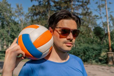 Dışarıda ağaçlarla çevrili bir parkta mavi tişörtlü ve güneş gözlüklü esmer bir adam voleybol maçından önce sokağın ortasında topla poz verirken ısınan bir voleybol topu dolduruyor.