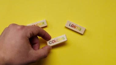 Gölgeleri olmayan sarı, sıcak bir arka plan. Erkek eli renkli kelimelerle ahşap küpler döşedi. Renklerin cinsiyeti LGBTQ.