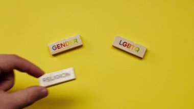 Gölgeleri olmayan sarı, sıcak bir arka plan. Erkek eli, siyah kelimelerle bezenmiş ahşap küpler serer. Din, LGBTQ cinsiyetini ortaya koyar.