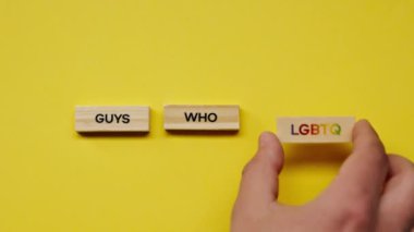 Gölgeleri olmayan sarı, sıcak bir arka plan. Erkek eli, LGBT elemanlarına renkli kelimeler içeren ahşap küpler yerleştirdi.