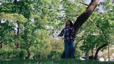 Dışarıda bir parkta ağaçların arasında teslim olma zeminine karşı esmer bir kız raket hareketleriyle servis atıyor.