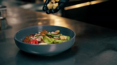Profesyonel bir mutfakta yakın plan çekimde şef krakerleri salata tabağı desenli bir kaseye kuru ekmek koyar.