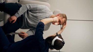 Klinikte genç bir doktorun dikey videosu. Kulak tıkayıcı. Hastanın burnunu ışıklarla muayene ediyor.