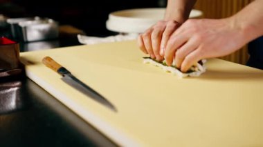 Japon lokantasında mavi üniformalı bir şef sarı bir tahtanın üzerinde balık sarması çeviriyor.