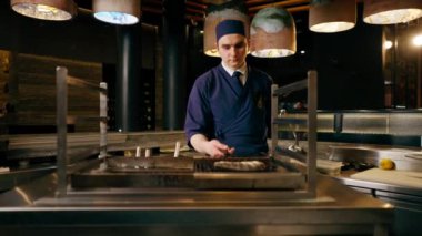 Bir Japon restoranında mavi üniformalı bir şef mutfaktaki ızgarada balık kızartıyor.