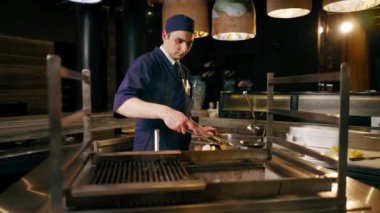 Bir Japon restoranında mavi üniformalı bir şef mutfaktaki ızgarada balık kızartıyor.
