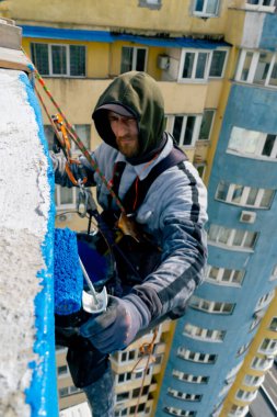 Çok renkli parlak evlerin arka planına karşı yüksek bir binanın çatısında, güvenlik ağı üzerine asılmış bir ressam duvarı maviye boyar.