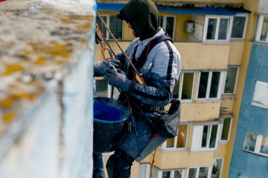 Çok renkli parlak evlerin arka planına karşı yüksek bir binanın çatısında, güvenlik ağı üzerine asılmış bir ressam duvarı maviye boyar.