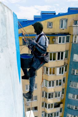 Çok renkli parlak evlerin arka planına karşı yüksek bir binanın çatısında, güvenlik ağı üzerine asılmış bir ressam duvarı maviye boyar.