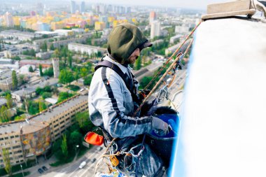 Çok renkli parlak evlerin arka planına karşı yüksek bir binanın çatısında, güvenlik ağı üzerine asılmış bir ressam duvarı maviye boyar.