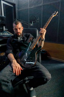 Kayıt odasındaki ekipmanda sakallı ve dövmeli yakışıklı bir adam kameraya gitar ile poz veriyor.