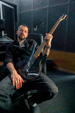 Kayıt odasındaki ekipmanda sakallı ve dövmeli yakışıklı bir adam kameraya gitar ile poz veriyor.