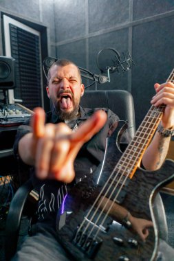 Kayıt odasının ekipmanında sakallı ve dövmeli yakışıklı bir adam kameraya gitar ile poz veriyor.