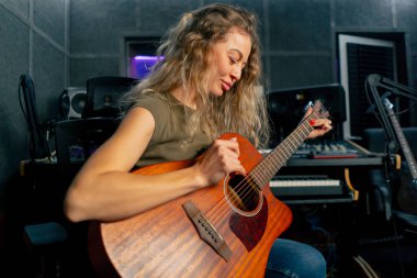 Güzel bir kıvırcık kadın, kayıt odasının ekipmanında turuncu akustik gitar çalmayı öğreniyor. Profesyonel bir eğitim.