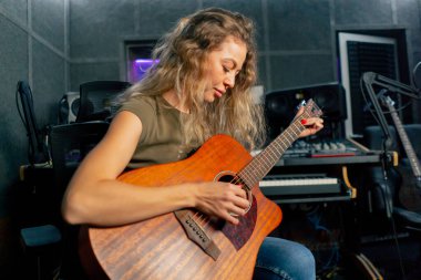 Güzel bir kıvırcık kadın, kayıt odasının ekipmanında turuncu akustik gitar çalmayı öğreniyor. Profesyonel bir eğitim.