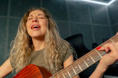 Güzel bir kıvırcık kadın, kayıt odasının ekipmanında turuncu akustik gitar çalmayı öğreniyor.