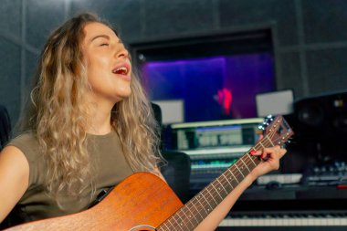 Güzel bir kıvırcık kadın, kayıt odasının ekipmanında turuncu akustik gitar çalmayı öğreniyor.