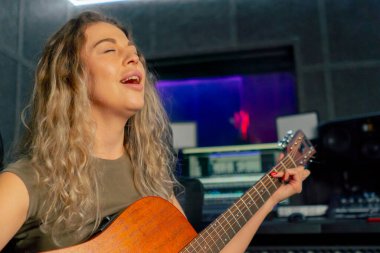 Güzel bir kıvırcık kadın, kayıt odasının ekipmanında turuncu akustik gitar çalmayı öğreniyor.