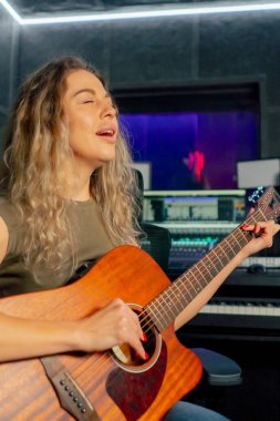 Güzel bir kıvırcık kadın, kayıt odasının ekipmanında turuncu akustik gitar çalmayı öğreniyor.