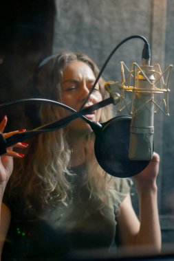 Bir kayıt stüdyosunda, temiz bir kayıt odasında, kulaklıklı güzel bir kıvırcık kız mikrofona şarkı söylüyor.