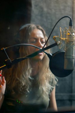 Bir kayıt stüdyosunda, temiz bir kayıt odasında, kulaklıklı güzel bir kıvırcık kız mikrofona şarkı söylüyor.