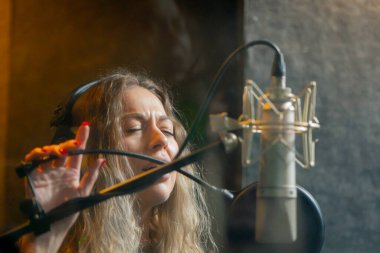 Bir kayıt stüdyosunda, temiz bir kayıt odasında, kulaklıklı güzel bir kıvırcık kız mikrofona şarkı söylüyor.