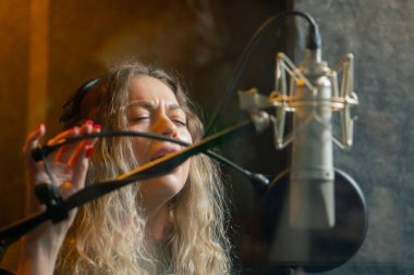 Bir kayıt stüdyosunda, temiz bir kayıt odasında, kulaklıklı güzel bir kıvırcık kız mikrofona şarkı söylüyor.