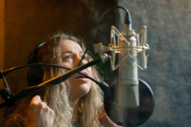 Bir kayıt stüdyosunda, temiz bir kayıt odasında, kulaklıklı güzel bir kıvırcık kız mikrofona şarkı söylüyor.