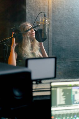 Bir kayıt stüdyosunda, temiz bir kayıt odasında, kulaklıklı güzel bir kıvırcık kız mikrofona şarkı söylüyor.