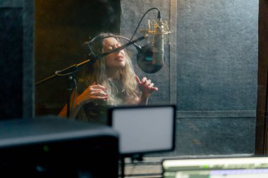 Bir kayıt stüdyosunda, temiz bir kayıt odasında, kulaklıklı güzel bir kıvırcık kız mikrofona şarkı söylüyor.