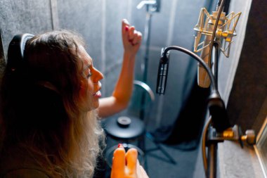 Bir kayıt stüdyosunda, temiz bir kayıt odasında, kulaklıklı güzel bir kıvırcık kız mikrofona şarkı söylüyor.