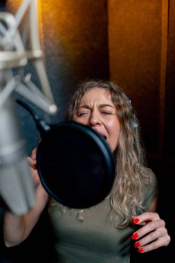 Bir kayıt stüdyosunda, temiz bir kayıt odasında, kulaklıklı güzel bir kıvırcık kız bir mikrofona şarkı söylüyor.