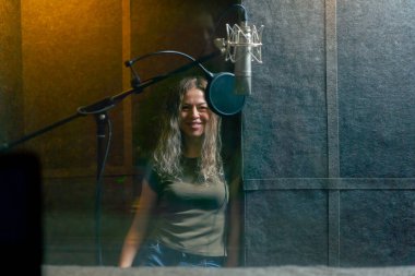 Bir kayıt stüdyosunda, temiz ses odasında, mikrofonun yanında duran kulaklıklı güzel bir kız kameraya bakıp gülümsüyor.