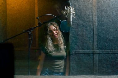 Bir kayıt stüdyosunda, temiz ses odasında, mikrofonun yanında duran kulaklıklı güzel bir kız kameraya bakıp gülümsüyor.