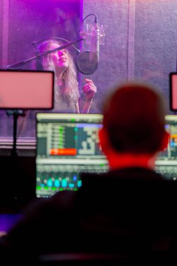 Bir kayıt stüdyosunda, temiz bir kayıt odasında kulaklıklı güzel bir kız mikrofona şarkı söylüyor. Şarkı bir monitörde kaydediliyor.