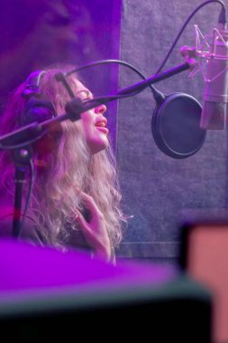 Kayıt stüdyosunda, temiz bir kayıt odasında kulaklıklı güzel bir kıvırcık kız menekşe ışıklarıyla mikrofona şarkı söylüyor.