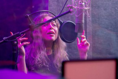 Kayıt stüdyosunda, temiz bir kayıt odasında kulaklıklı güzel bir kıvırcık kız menekşe ışıklarıyla mikrofona şarkı söylüyor.