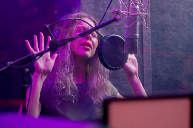 Kayıt stüdyosunda, temiz bir kayıt odasında kulaklıklı güzel bir kıvırcık kız menekşe ışıklarıyla mikrofona şarkı söylüyor.