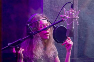 Kayıt stüdyosunda, temiz bir kayıt odasında kulaklıklı güzel bir kıvırcık kız menekşe ışıklarıyla mikrofona şarkı söylüyor.