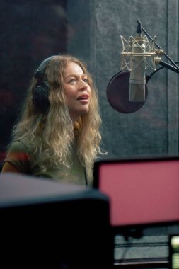 Bir kayıt stüdyosunda, temiz bir kayıt odasında, kulaklıklı güzel bir kıvırcık kız bir mikrofona şarkı söylüyor.