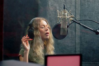 Bir kayıt stüdyosunda, temiz bir kayıt odasında, kulaklıklı güzel bir kıvırcık kız bir mikrofona şarkı söylüyor.