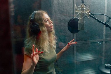 Bir kayıt stüdyosunda, temiz bir kayıt odasında, kulaklıklı güzel bir kıvırcık kız bir mikrofona şarkı söylüyor.