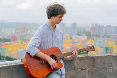 Renkli parlak evlerin arkaplanına karşı yüksek bir binanın çatısında yakışıklı bir genç adam duruyor ve akustik gitar çalıyor.