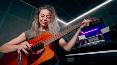 Güzel bir kıvırcık kadın, kayıt odasının ekipmanında turuncu akustik gitar çalmayı öğreniyor. Profesyonel bir eğitim.