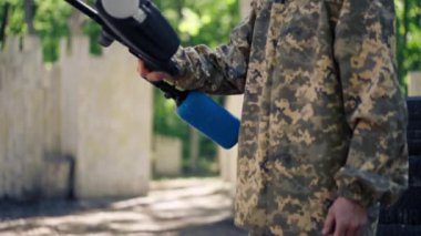 Vahşi doğada ağaçların yanında pintball üniformalı bir oyuncu mavi balon takım kıyafetleriyle bir tabanca tutuyor.